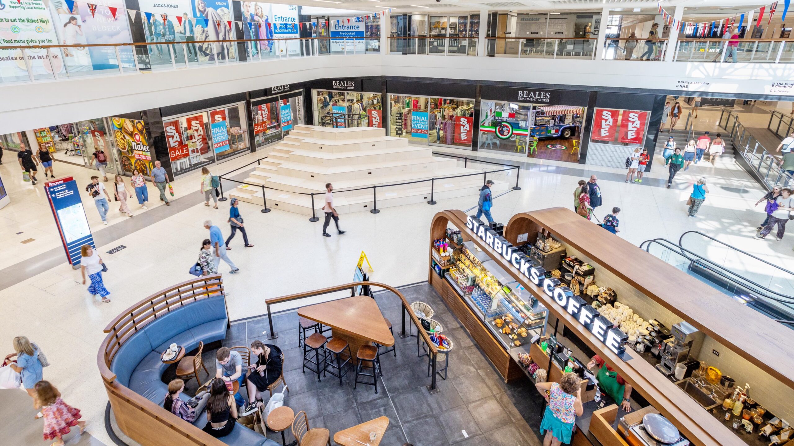 Image of The Dolphin Poole interior: A bright and spacious indoor shopping center with multiple levels. In the foreground, a Starbucks Coffee kiosk with patrons seated around it is visible. Shoppers are walking along the wide walkways on both levels, with various stores including Beales displaying large sale signs. An open stairway and escalator are seen on the right, facilitating movement between floors. Colorful banners and advertisements add to the lively atmosphere. The scene captures a mix of people shopping, socializing, and enjoying their time in the mall.