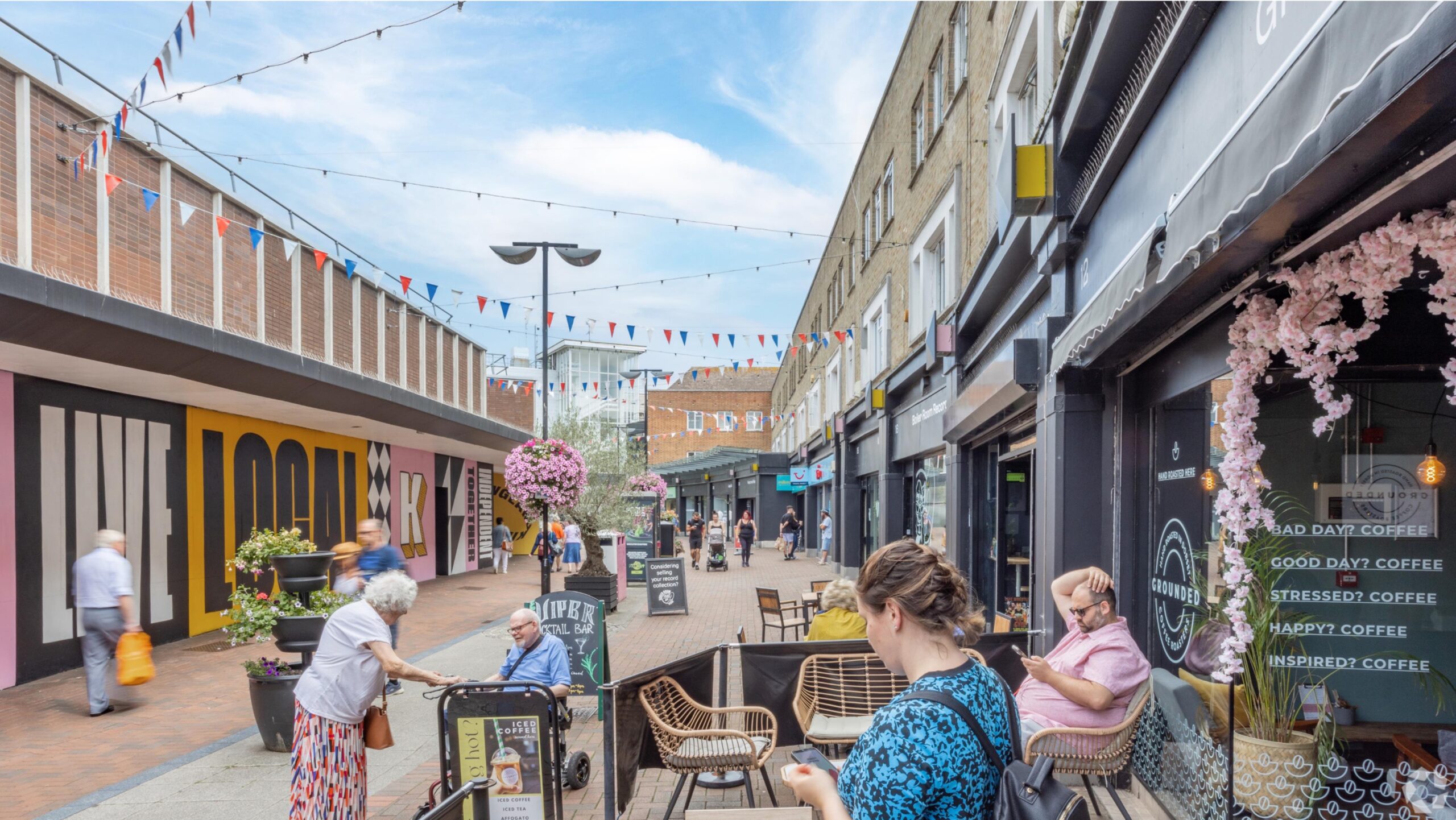 A lively street scene at Kingland Crescent, Poole, featuring a pedestrian area lined with shops and outdoor seating. People are enjoying the pleasant weather, with some sitting at tables outside a café named 